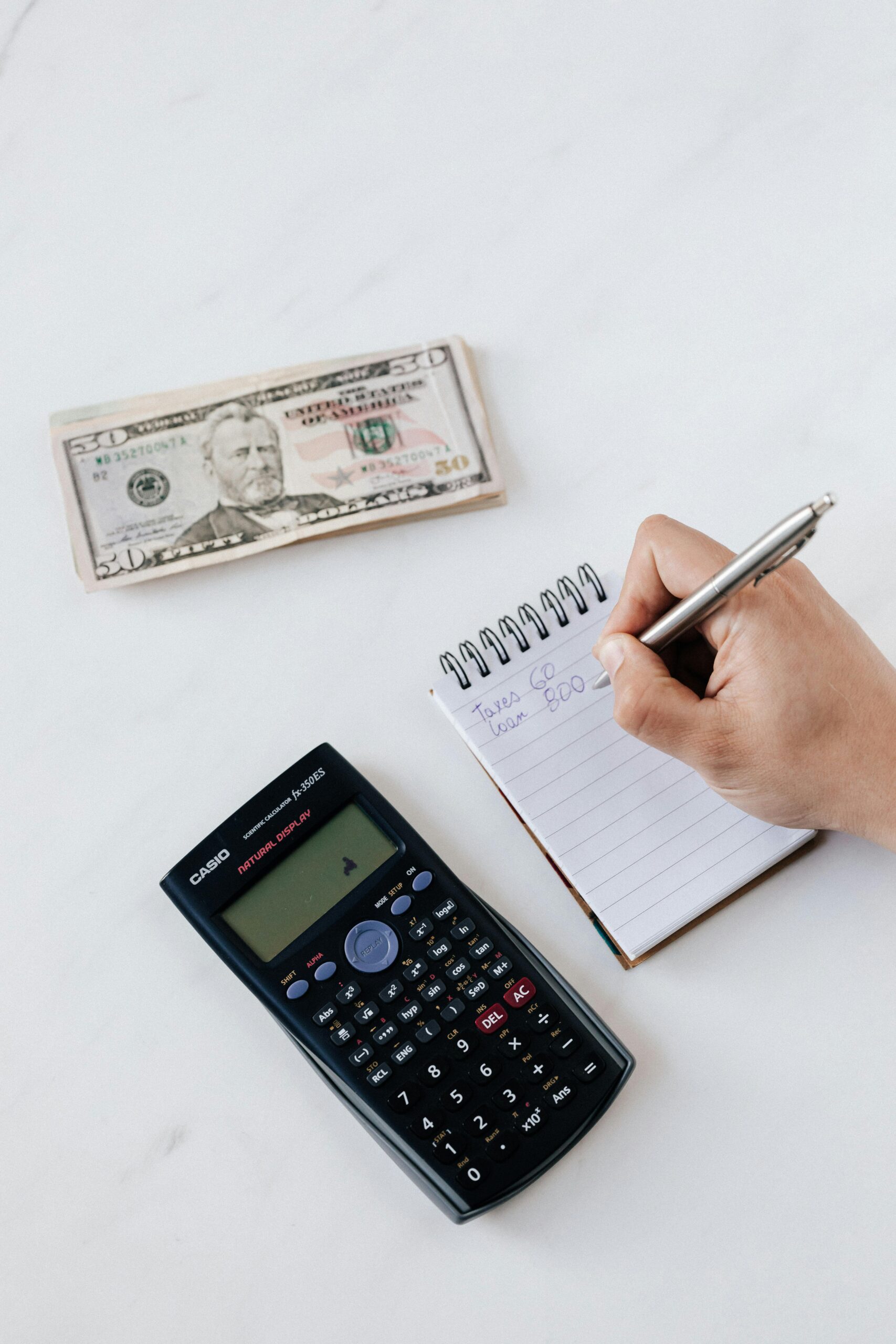 Close-up of hand writing in a notepad with a calculator and dollar bill on a desk, focusing on budgeting.